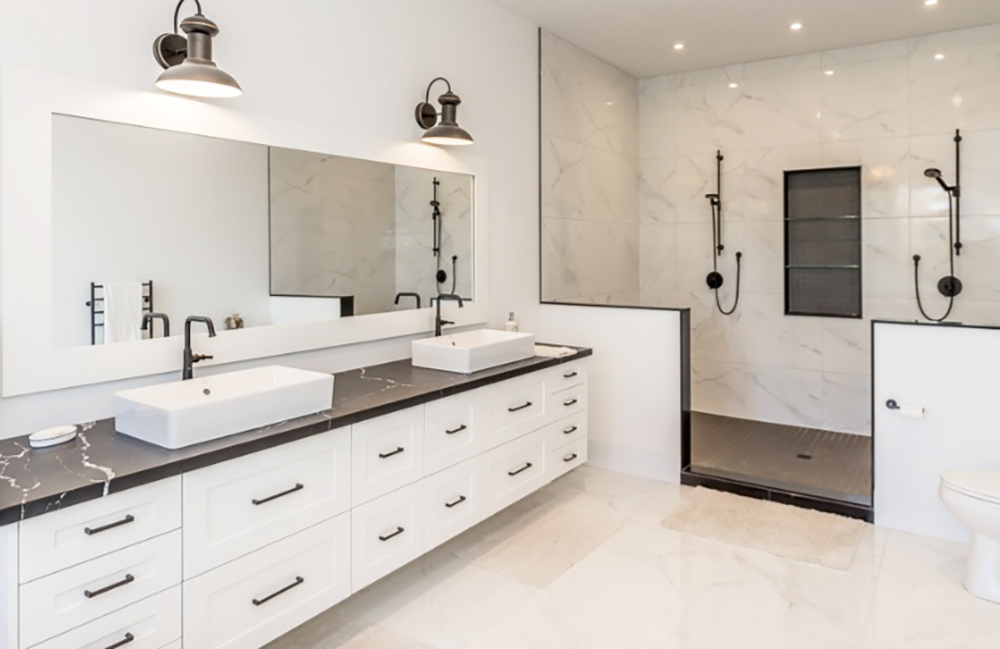 A bathroom renovation with two rectangular white sinks, black faucets, marble counter top, two overhead lights and a mirror, as well as a walk in shower